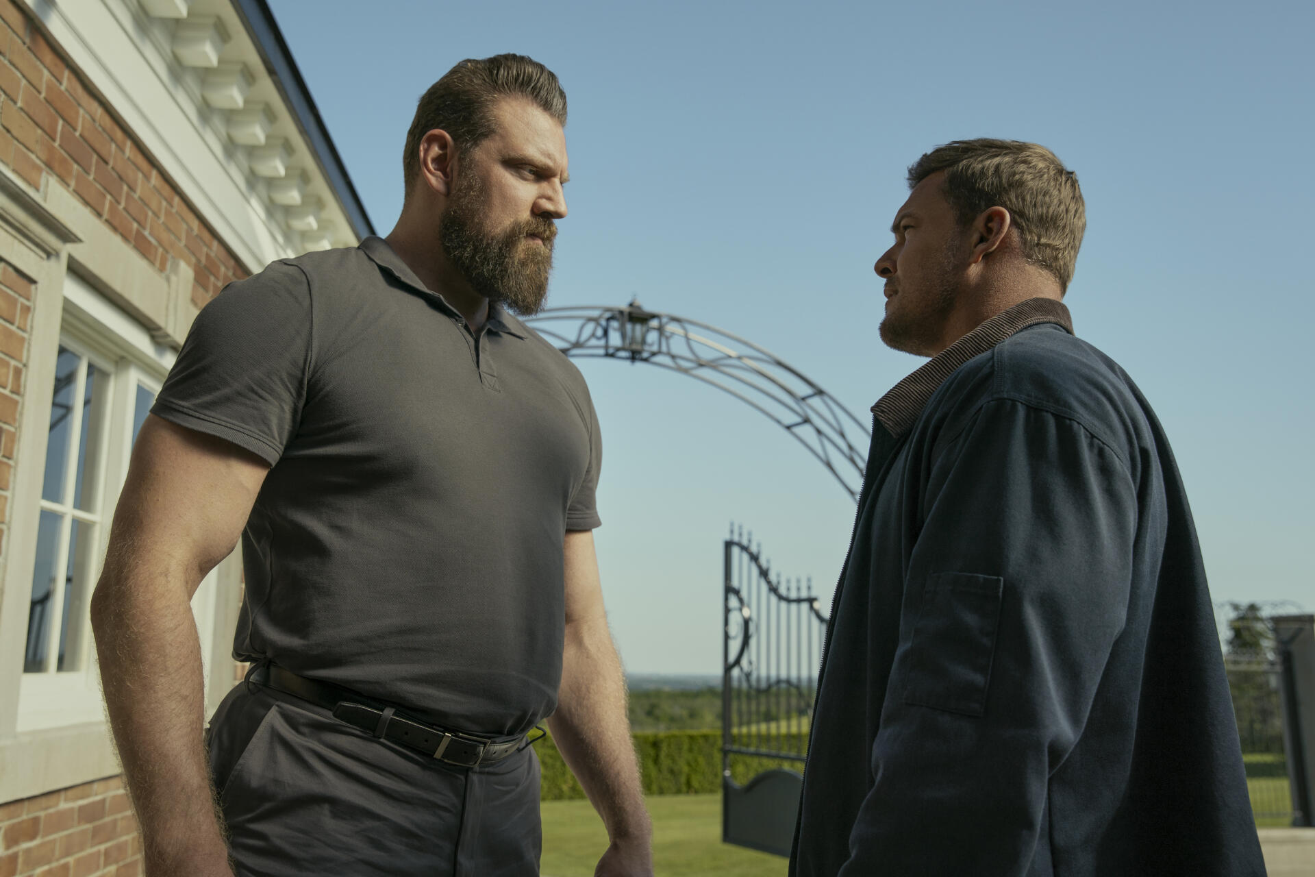 HD desktop wallpaper featuring Alan Ritchson and Olivier Richters from the TV show Reacher, standing face-to-face outdoors near a gated entrance.