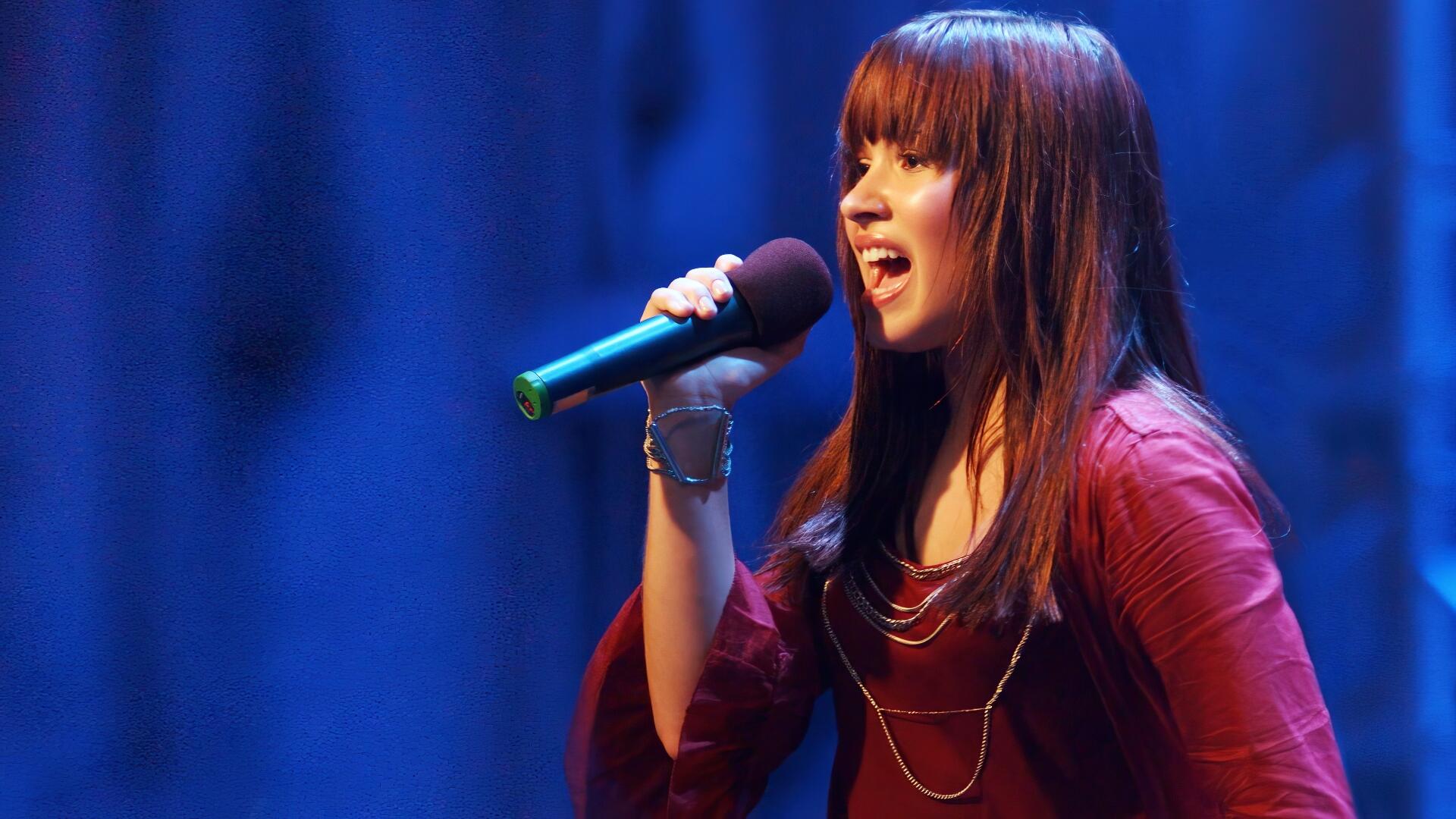 HD desktop wallpaper showing a Camp Rock singer mid-song, holding a microphone onstage with blue curtains behind her.