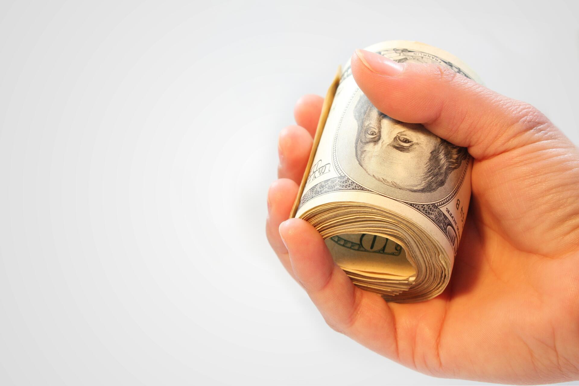 Close-up of a hand gripping a rolled stack of US dollar banknotes on a white background — HD PC desktop wallpaper.