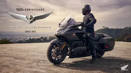 A Honda GL 1800 Gold Wing motorcycle stands proudly with a rider in gear, set against a scenic backdrop, celebrating its 50th anniversary since 1975.