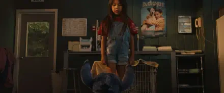 A scene from the movie Lilo & Stitch (2025) shows a young girl in overalls standing over a blue stuffed creature in a room with animal cages and an adoption poster on the wall.