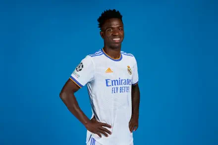 Vinícius Júnior smiles confidently in his Real Madrid C.F. soccer uniform, set against a vibrant blue background. This high-definition image captures his dynamic presence in sports.