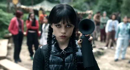 HD desktop wallpaper of Wednesday from the TV show Wednesday — pale, braided girl with dark bangs staring defiantly while holding a megaphone at an outdoor fair.