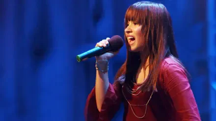 HD desktop wallpaper showing a Camp Rock singer mid-song, holding a microphone onstage with blue curtains behind her.