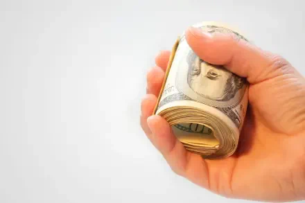 Close-up of a hand gripping a rolled stack of US dollar banknotes on a white background — HD PC desktop wallpaper.