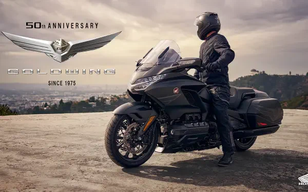 A Honda GL 1800 Gold Wing motorcycle stands proudly with a rider in gear, set against a scenic backdrop, celebrating its 50th anniversary since 1975.
