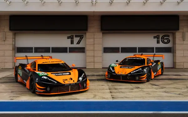 Two McLaren GT3 EVO race cars in bright orange and black are parked in front of garage doors numbered 16 and 17, set against a sleek, modern backdrop. A striking 4K Ultra HD image.