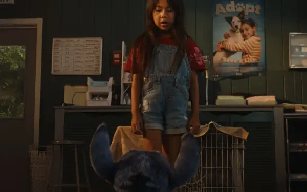 A scene from the movie Lilo & Stitch (2025) shows a young girl in overalls standing over a blue stuffed creature in a room with animal cages and an adoption poster on the wall.