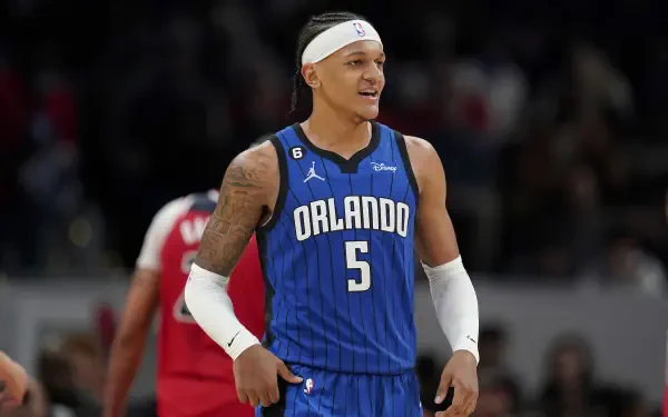 Orlando Magic player Paolo Banchero smiles on the court, wearing the team's blue jersey and headband, featured as a vibrant HD desktop wallpaper.