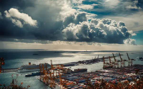 A 4K Ultra HD view of Barcelona's man-made Mediterranean port under dramatic clouds, showcasing shipping containers and cranes along the waterfront.
