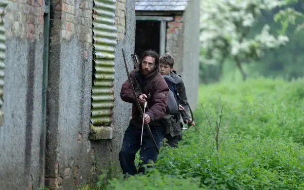 4K Ultra HD desktop wallpaper from the movie 28 Years Later, showing two armed men cautiously moving alongside an abandoned, overgrown building.