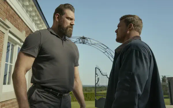 HD desktop wallpaper featuring Alan Ritchson and Olivier Richters from the TV show Reacher, standing face-to-face outdoors near a gated entrance.