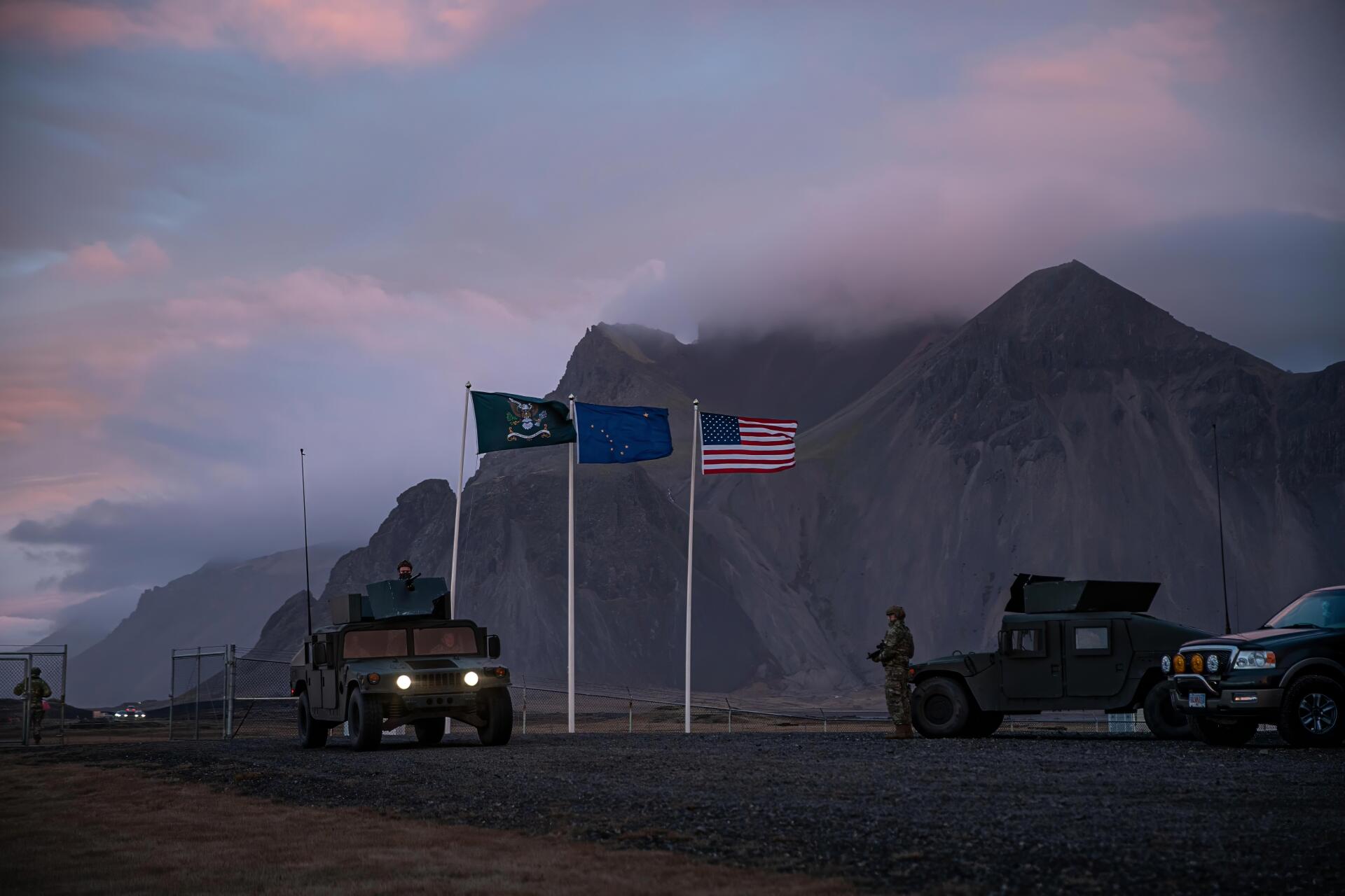 4K Ultra HD movie wallpaper from A House of Dynamite: military convoy and Humvees beside three flags at dusk against a rugged mountain backdrop, PC desktop background.