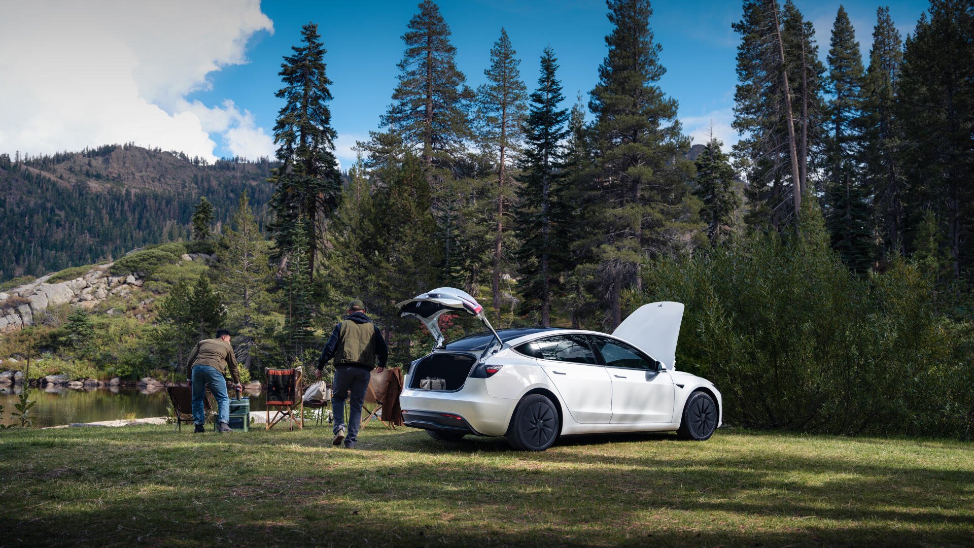 4K Ultra HD PC desktop wallpaper of a white Tesla parked at a forest campsite, trunk and hood open, people and camping gear beside a lake amid tall pines and mountain backdrop.