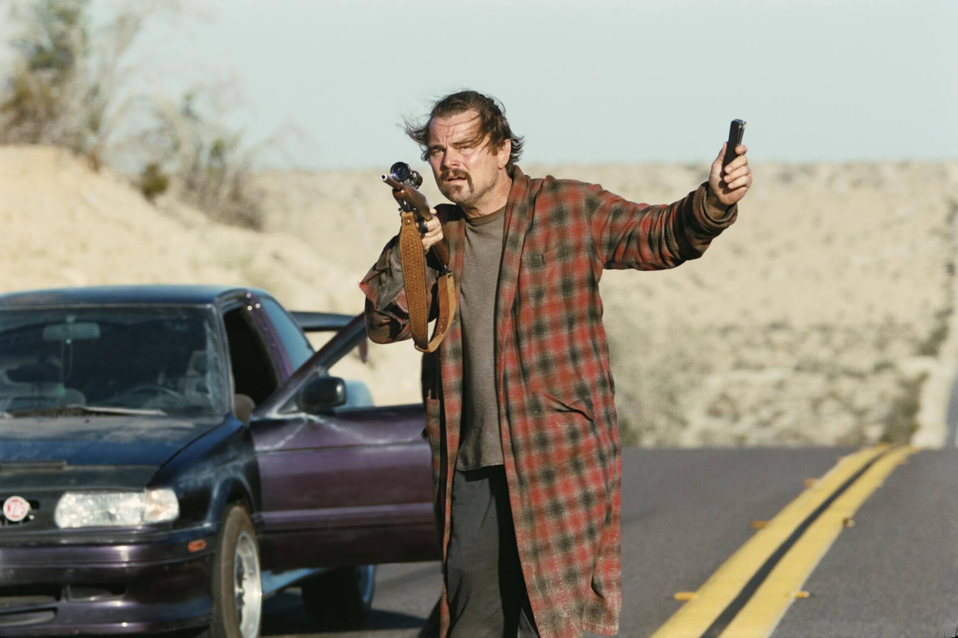 4K Ultra HD PC desktop wallpaper for the movie One Battle After Another: rugged male actor in a plaid coat aiming a rifle, holding a phone beside a car on a desert road.