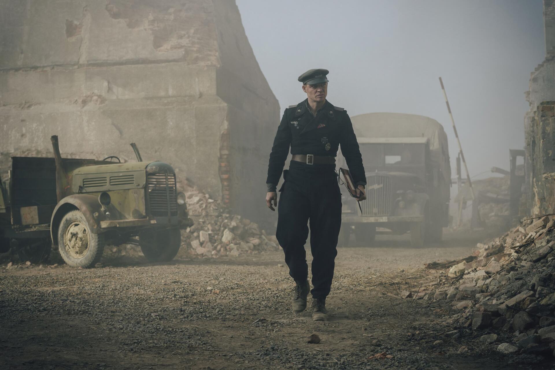 Der Tiger movie scene: a uniformed officer walks down a rubble-strewn street flanked by military trucks. 4K Ultra HD PC desktop wallpaper/background.