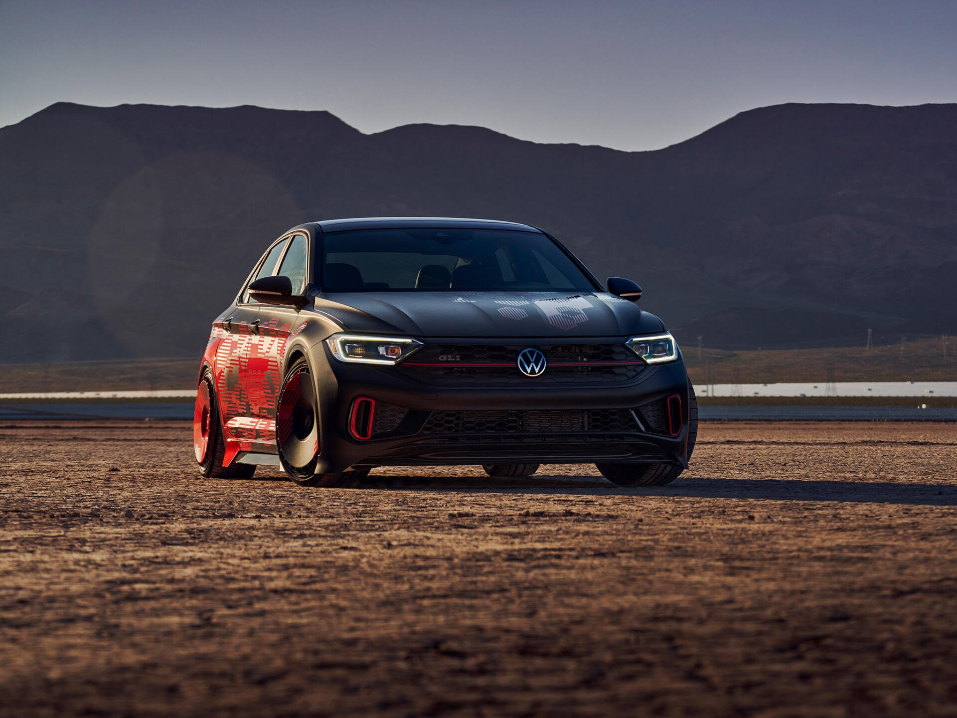 HD PC desktop wallpaper: vehicle — a black Volkswagen Jetta parked on a desert flat at dusk, low-angle view highlighting its sporty front end with mountains in the background.