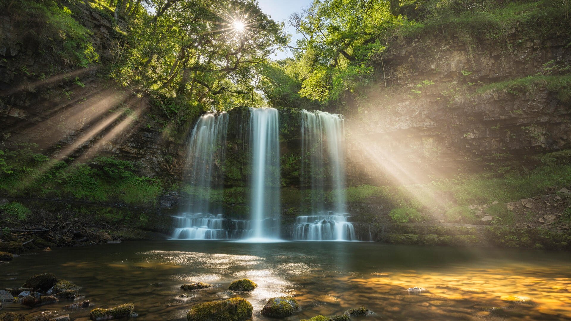 4K Ultra HD PC wallpaper of a Welsh waterfall: twin cascades through lush woodland with sunlight rays piercing the canopy and golden light on the pool — nature photography.