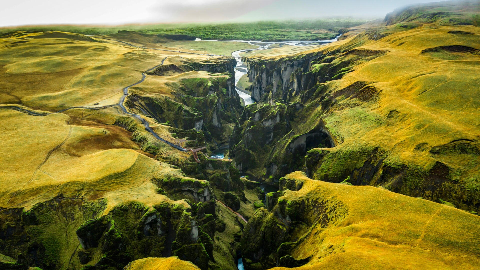 Aerial 5K Ultra HD wallpaper of Fjaðrárgljúfur canyon, Iceland: winding river through steep green cliffs and mossy plateaus under soft light.