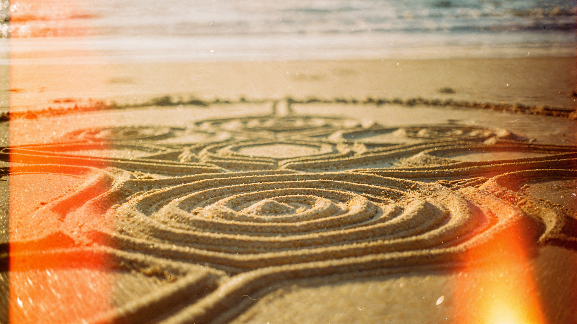 Close-up of intricate circular sand art etched in wet beach sand at sunrise, warm light leaks — 4K Ultra HD PC desktop wallpaper and background.