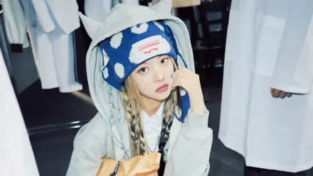 Young woman with braided hair in a blue polka-dot beanie and hooded jacket, holding a snack bag in a studio; K-pop, South Korean music aesthetic; 4K Ultra HD PC desktop wallpaper.