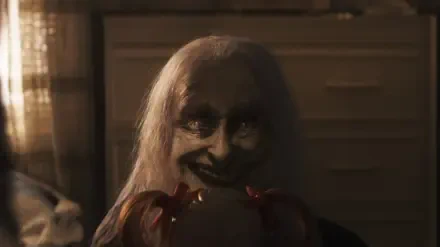 Creepy pale-haired demon with a sinister grin clutching a doll in a dim room — The Conjuring: Last Rites 2K Quad HD PC desktop wallpaper/background.