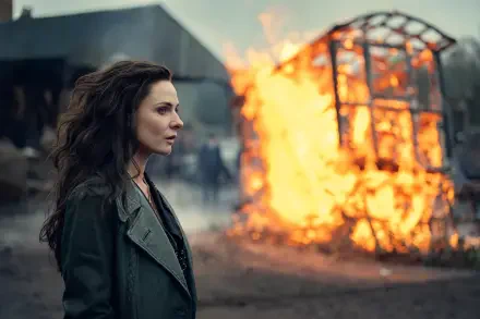 Lead actress in profile wearing a coat watches a massive burning wheel amid smoky ruins — cinematic shot from the movie Peaky Blinders: The Immortal Man, 8K Ultra HD desktop wallpaper.