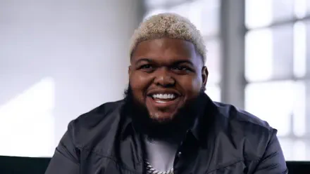 HD PC desktop wallpaper of a smiling male streamer with short bleached-blond hair and a beard, wearing a dark jacket against a soft-lit background.