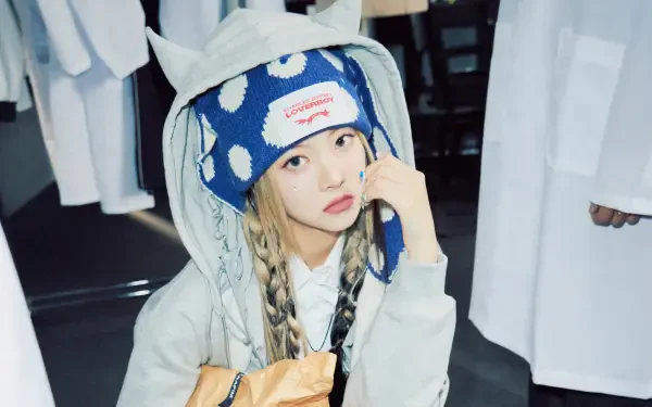 Young woman with braided hair in a blue polka-dot beanie and hooded jacket, holding a snack bag in a studio; K-pop, South Korean music aesthetic; 4K Ultra HD PC desktop wallpaper.