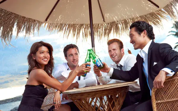 Hawaii Five-0 4K Ultra HD PC wallpaper: four smiling people in evening wear clinking bottles beneath a thatched beach umbrella at an oceanfront table.