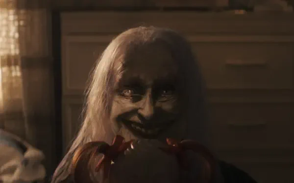 Creepy pale-haired demon with a sinister grin clutching a doll in a dim room — The Conjuring: Last Rites 2K Quad HD PC desktop wallpaper/background.