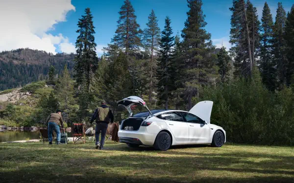 4K Ultra HD PC desktop wallpaper of a white Tesla parked at a forest campsite, trunk and hood open, people and camping gear beside a lake amid tall pines and mountain backdrop.
