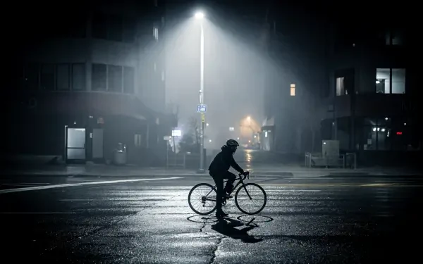 Silhouetted bicyclist crossing a wet, dimly lit urban street beneath a lone streetlight, pavement reflections shimmering — 4K Ultra HD PC desktop wallpaper and background.