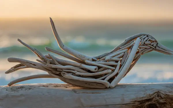 Driftwood sculpture shaped like a bird resting on a weathered log with soft ocean and pastel sunset bokeh — 4K Ultra HD PC Desktop Wallpaper and Background
