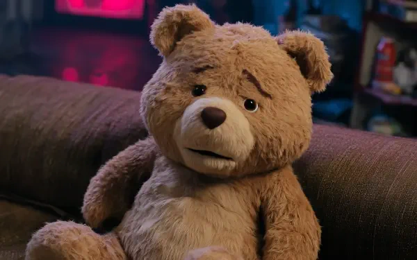 Close-up of Ted, the plush bear from the Ted TV show/movie, seated on a couch in a dim living room — 2K Quad HD PC desktop wallpaper/background.