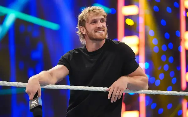 Blond man in a black T‑shirt leaning on WWE ring ropes, smiling under colorful arena lights — 2K Quad HD PC desktop wallpaper background.