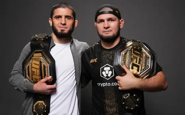 HD desktop wallpaper of two UFC champions posing shoulder-to-shoulder with gold title belts, one in a jacket and the other in a cap and black shirt, studio portrait on a gray background.