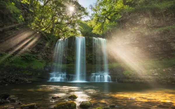 4K Ultra HD PC wallpaper of a Welsh waterfall: twin cascades through lush woodland with sunlight rays piercing the canopy and golden light on the pool — nature photography.