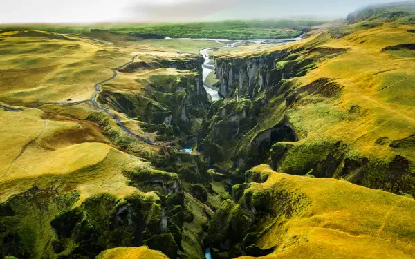 Aerial 5K Ultra HD wallpaper of Fjaðrárgljúfur canyon, Iceland: winding river through steep green cliffs and mossy plateaus under soft light.
