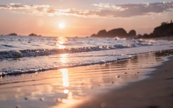 Beach sunset 4K Ultra HD PC desktop wallpaper and background: golden sun sinking behind distant rocks, gentle waves washing reflective wet sand beneath a pastel sky.
