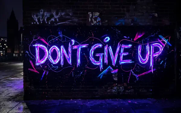 Neon DON'T GIVE UP graffiti in purple and blue glowing on a wet, dark brick wall with pavement reflections — 5K Ultra HD PC desktop wallpaper/background.