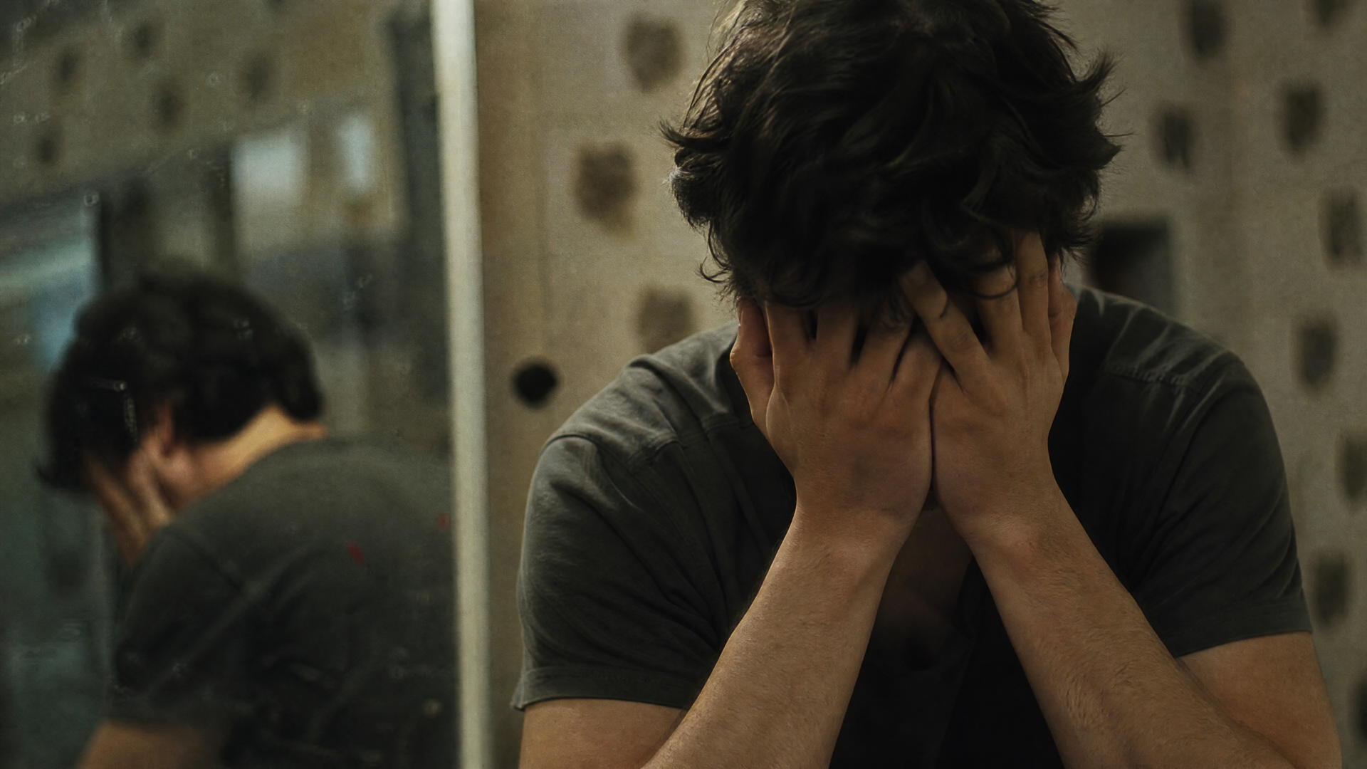 4K Ultra HD PC Desktop Wallpaper and Background: moody low-light scene of a seated man with hands covering his face, mirror reflection behind him, muted, somber tones.