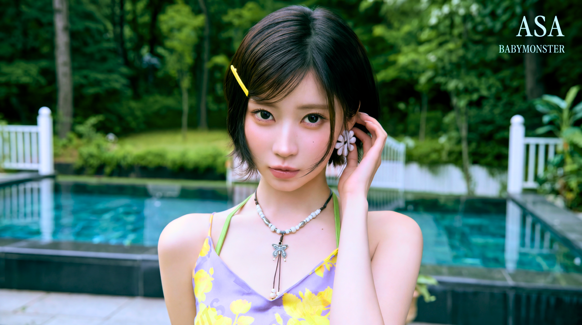 Young woman in a yellow floral dress by a pool, short dark hair with a hairpin, layered necklaces, lush green trees in background — 5K Ultra HD PC desktop wallpaper.