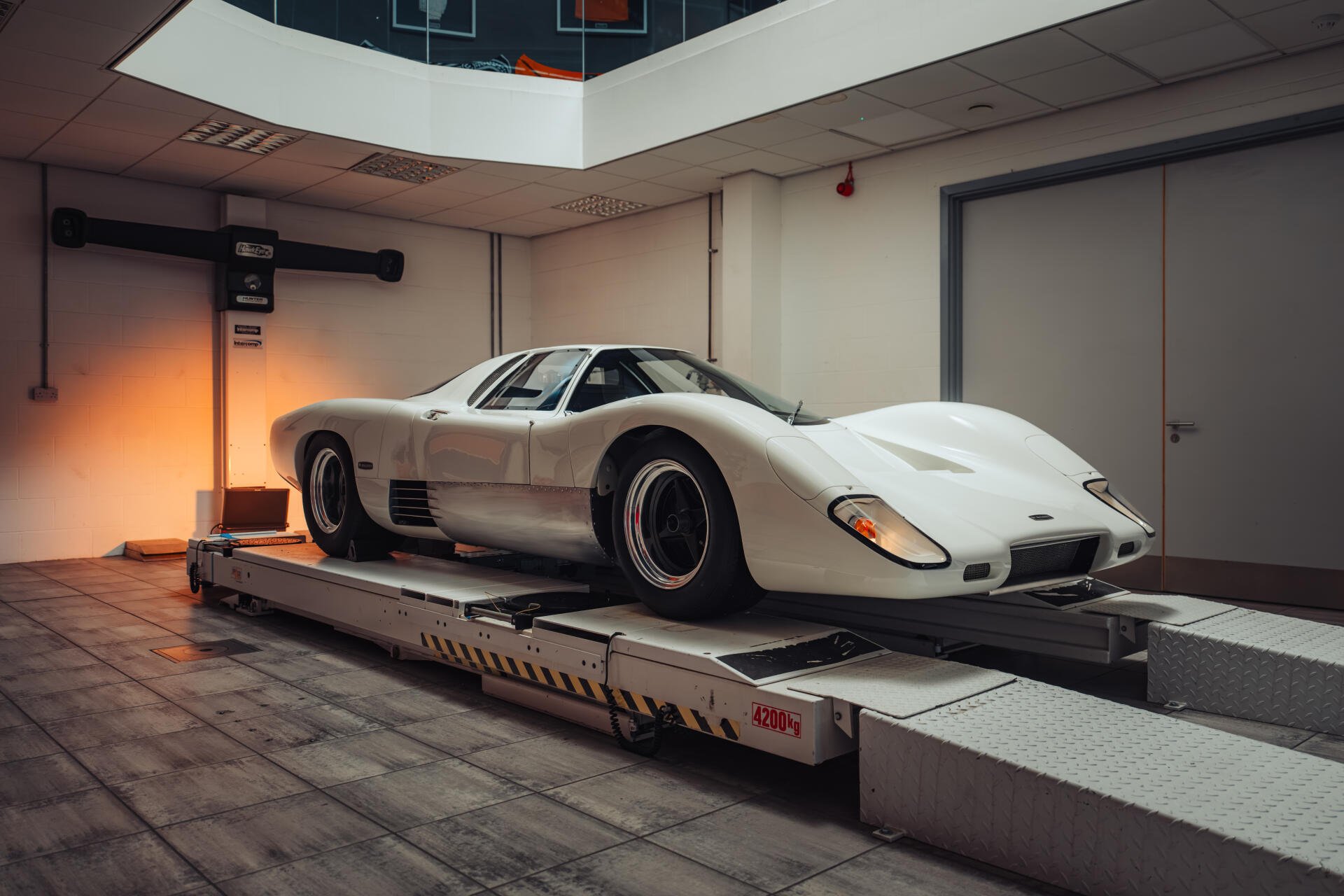 8K Ultra HD PC desktop wallpaper of a white McLaren G6GT sports car on a workshop lift, showcasing its low-slung profile and vintage racing wheels.