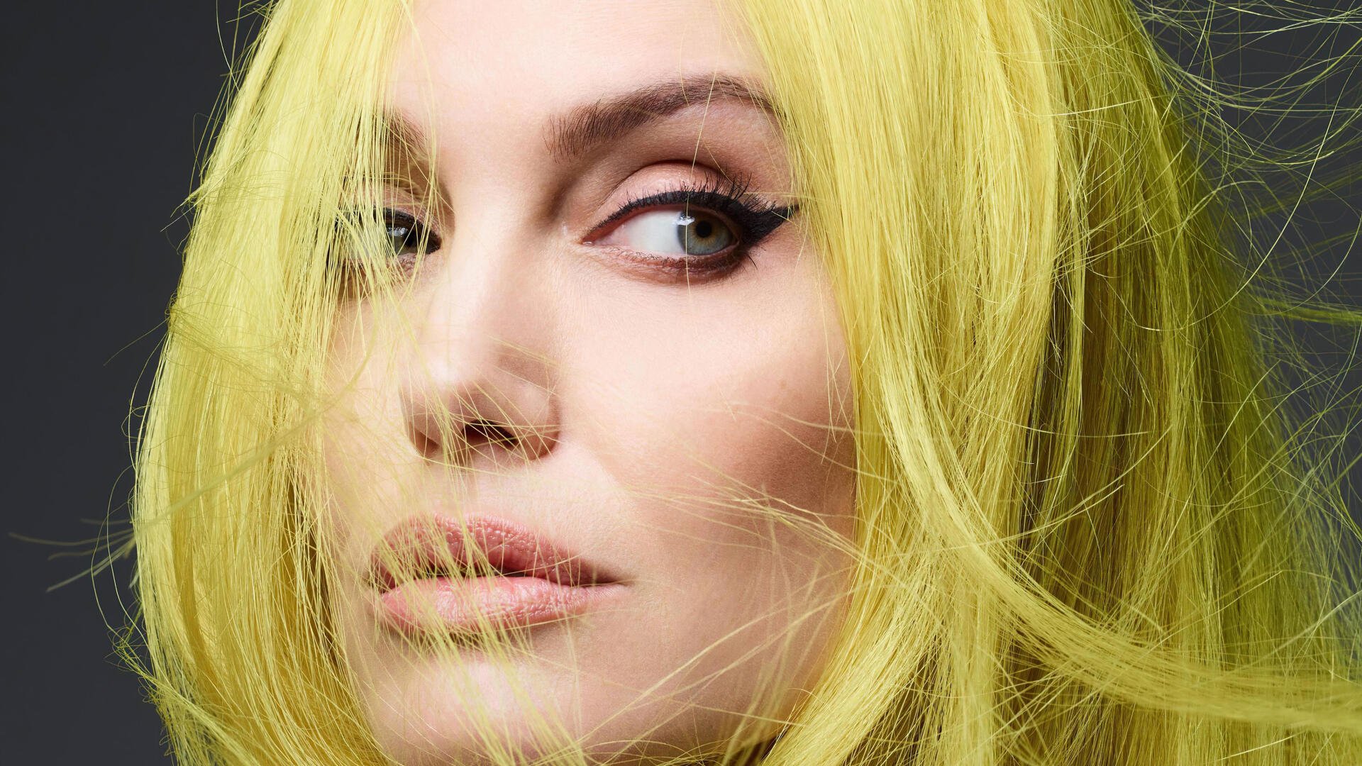 Close-up portrait of a woman with vivid yellow hair and winged eyeliner, soft skin tones, gazing off-frame — 4K Ultra HD PC desktop wallpaper/background.