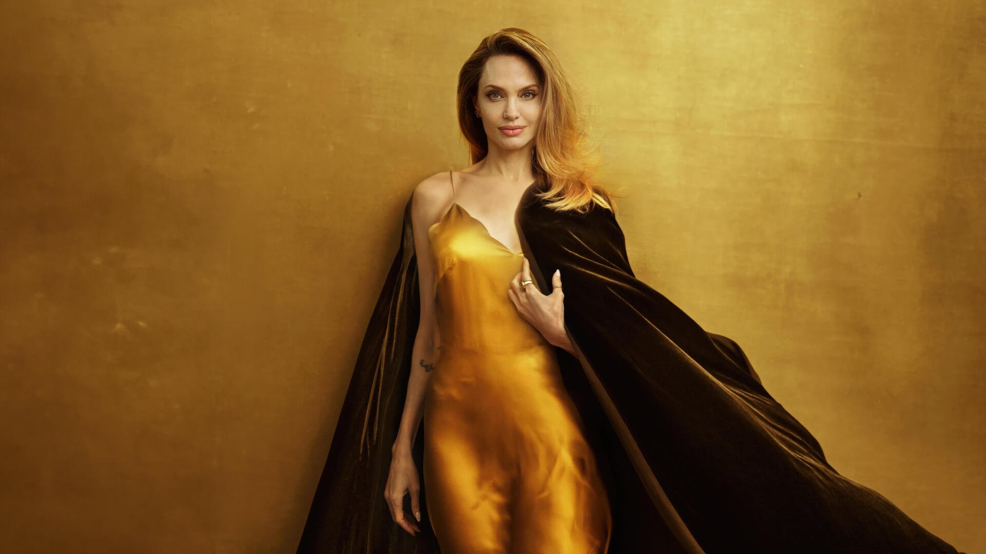 Glamorous portrait of a woman in a golden satin gown and dark cloak against a warm gold backdrop, 4K Ultra HD PC desktop wallpaper.