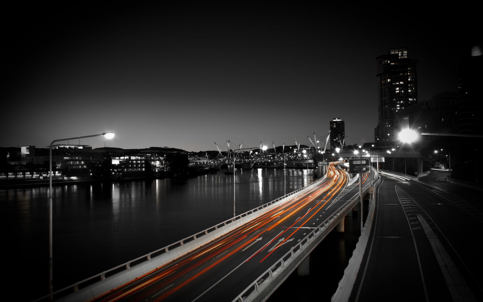 Nighttime cityscape featuring a man-made road with streaks of traffic lights along a waterfront, presented in high-definition for a PC desktop wallpaper background.