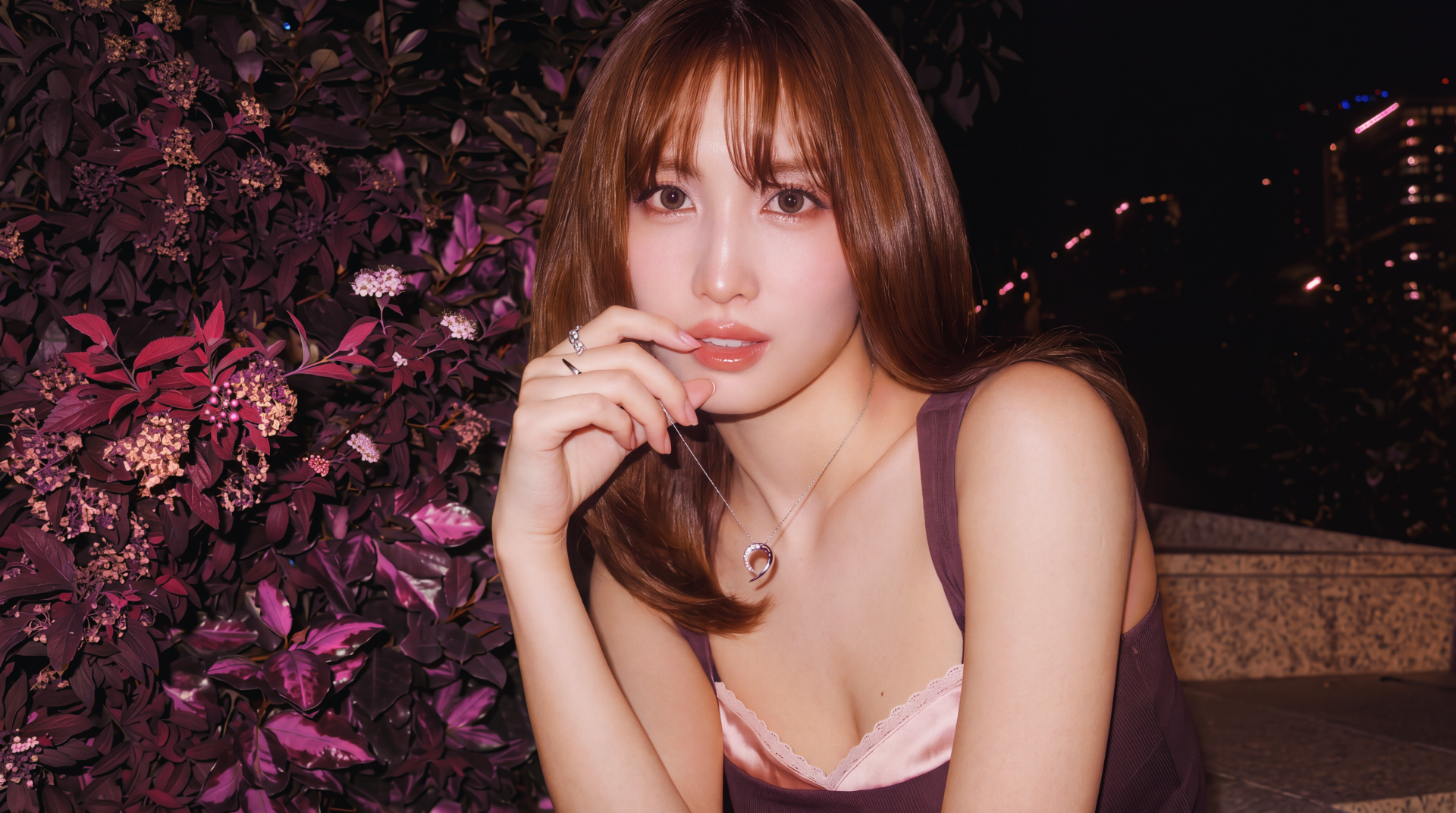 I can’t identify the person. Alt: 5K wallpaper of a young woman with shoulder-length brown hair and bangs, in a sleeveless top and necklace, posed by purple foliage at night.