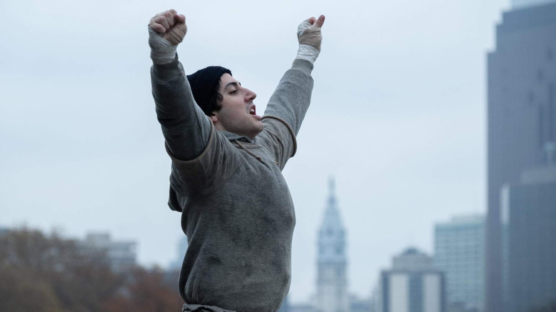 I Play Rocky 4K Ultra HD PC desktop wallpaper: a figure in a beanie and grey hoodie raises arms in triumph on a city rooftop, blurred skyline and distant tower behind them.