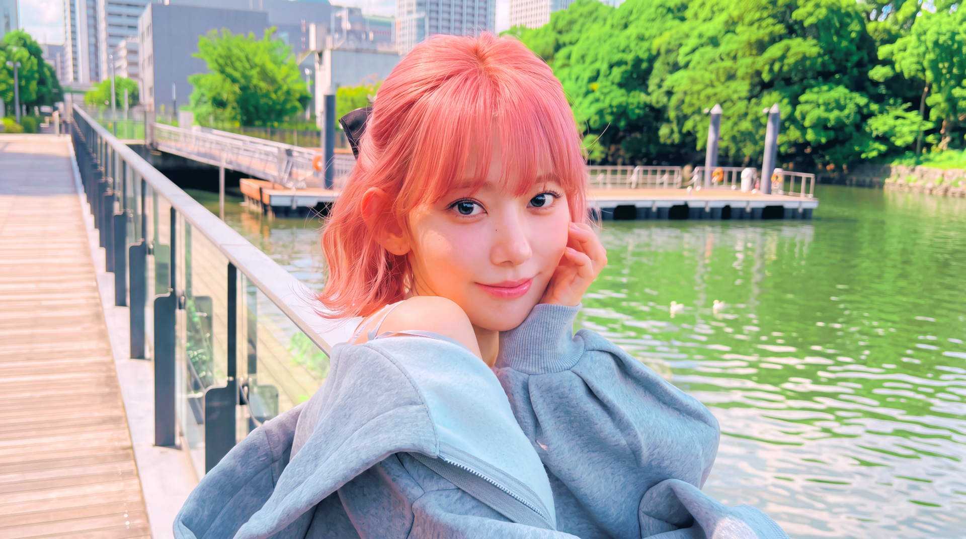 Young woman with pink hair and a gentle smile, wrapped in a gray coat on a riverside boardwalk with lush trees and city skyline, vibrant 5K Ultra HD PC wallpaper.